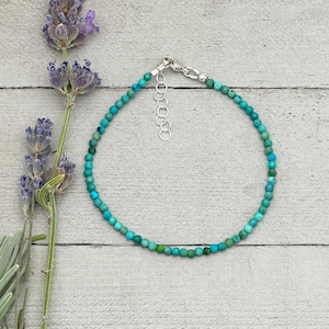May include: A turquoise beaded bracelet with a silver clasp and extender chain. The bracelet is made of small, round, turquoise beads. The background is a light gray wooden surface with sprigs of lavender.