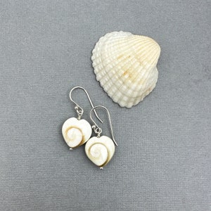 May include: Heart-shaped earrings with a spiral design in cream and brown, hanging from silver hooks. A white seashell with a textured surface is next to the earrings on a gray background. The earrings are a unique accessory.