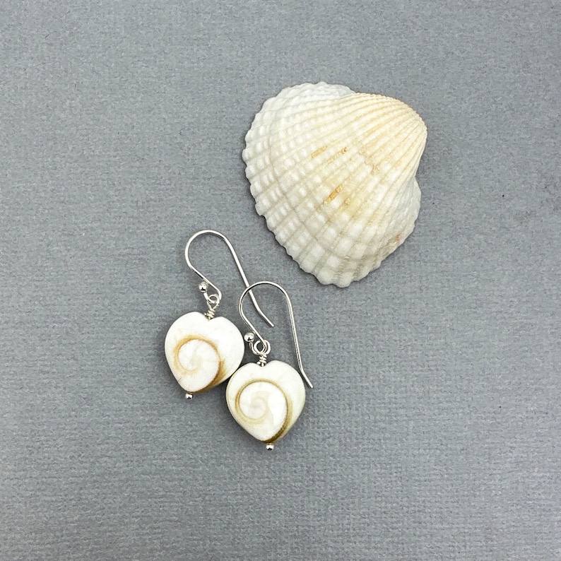 May include: Heart-shaped earrings with a spiral design in cream and brown, hanging from silver hooks. A white seashell with a textured surface is next to the earrings on a gray background. The earrings are a unique accessory.