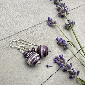 May include: A pair of silver dangle earrings with purple and white striped glass beads. The earrings are photographed against a light grey wooden background with lavender flowers.