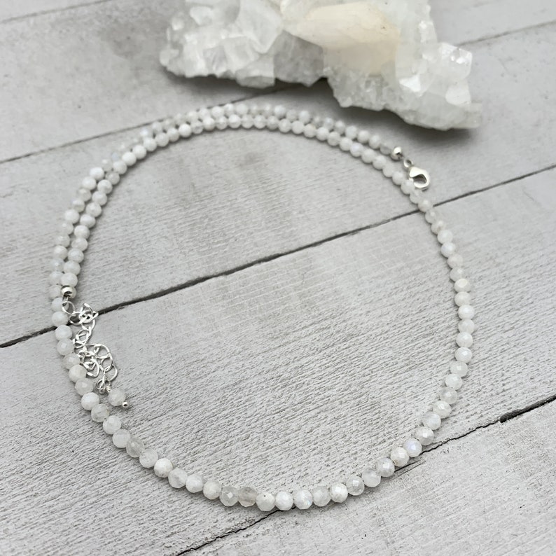 Puede incluir: Un collar de piedra lunar blanca con un cierre plateado. El collar est&aacute; hecho de peque&ntilde;as cuentas redondas y est&aacute; colocado sobre una superficie de madera blanca.