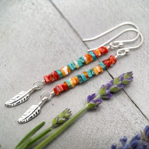 May include: Handmade earrings featuring colorful, square-cut beads in shades of red, orange, turquoise, and white. Each earring has a silver feather charm and a silver hook. The earrings are displayed with a sprig of lavender.