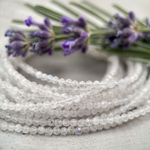 Puede incluir: Una hebra de cuentas de piedra lunar blanca, con un sutil brillo iridiscente, se muestra sobre una superficie blanca. Las cuentas son peque&ntilde;as y redondas, y est&aacute;n dispuestas en una espiral suelta. Una ramita de lavanda se coloca encima de las cuentas, a&ntilde;adiendo un toque de color y fragancia.