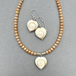 May include: A necklace and earring set featuring heart-shaped pendants and earrings with a spiral design. The necklace and earrings are paired with a strand of peach-colored pearls. The jewelry is set against a gray background.