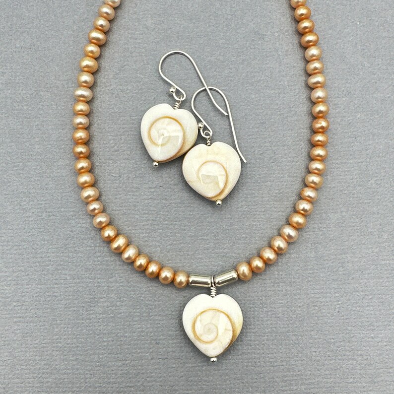 May include: A necklace and earring set featuring heart-shaped pendants and earrings with a spiral design. The necklace and earrings are paired with a strand of peach-colored pearls. The jewelry is set against a gray background.