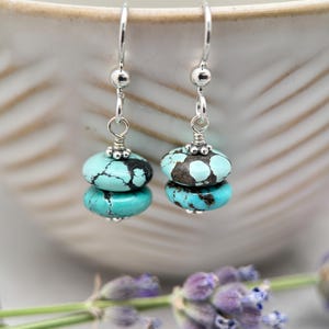 May include: A pair of turquoise and silver earrings. Each earring features two turquoise beads with dark brown veining, stacked and connected by silver wire and a silver hoop. The earrings are displayed against a blurred background.
