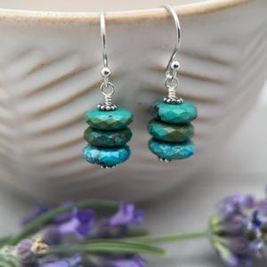 May include: A pair of dangle earrings featuring three stacked, faceted turquoise-colored beads on each silver-toned hook. The beads display natural variations in blue and green tones with dark matrix inclusions, suspended from simple silver wire findings.