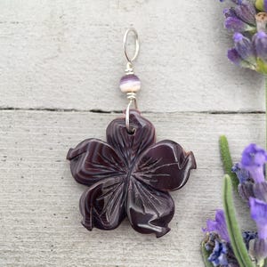 Op de afbeelding: Een donkerpaarse, vijfbladige bloemhanger gesneden uit steen met zichtbare gelaagde banden. De hanger is bevestigd aan een zilverkleurige metalen ring en heeft een kleine, ronde, gestreepte kraal in wit- en paarstinten boven de bloem.