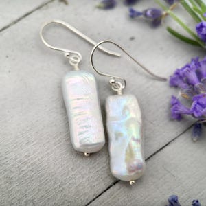 May include: A pair of white, rectangular pearl earrings with silver hooks. The pearls have an iridescent sheen, reflecting colors of blue, green, and yellow. The earrings are displayed on a light gray wooden surface, with purple flowers in the background.