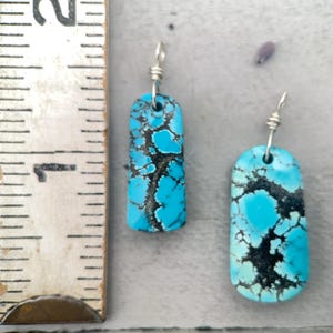 May include: Two turquoise earrings with a rectangular shape and a silver wire hook. The earrings have a black matrix pattern throughout the turquoise stone. The earrings are displayed on a light grey surface.