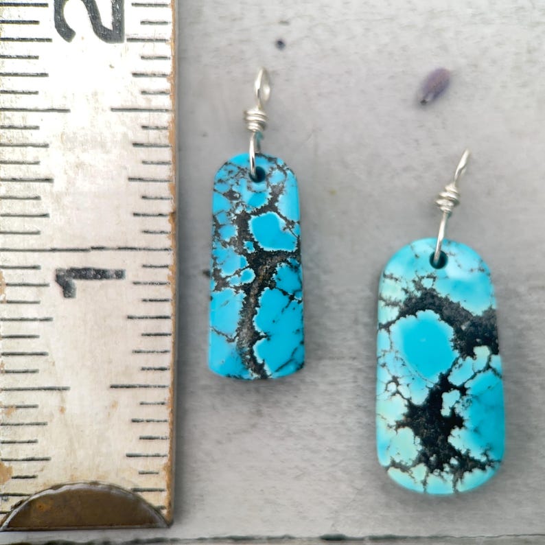 May include: Two turquoise earrings with a rectangular shape and a silver wire hook. The earrings have a black matrix pattern throughout the turquoise stone. The earrings are displayed on a light grey surface.