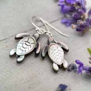 Puede incluir: Pendientes de tortuga plateados. Cada pendiente presenta un diseño de tortuga con una concha de nácar y aletas y cabeza de color gris oscuro. Los pendientes están sobre una superficie gris claro con flores moradas.