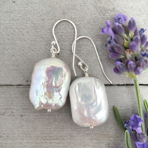 Pode incluir: Um par de brincos pendentes com grandes pérolas barrocas brancas e iridescentes, suspensas por ganchos de metal prateado simples. As pérolas exibem um brilho lustroso e multicolorido com tons subtis de rosa e azul numa superfície natural.