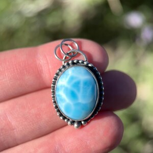 Puede incluir: Un colgante de forma ovalada con una piedra lisa de color azul claro y patrones veteados en blanco. La piedra est&aacute; engastada en un marco de metal plateado con un borde de cuentas y una anilla circular en la parte superior para colgar.