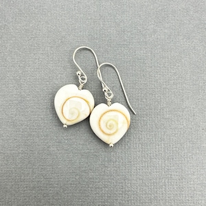 May include: A pair of heart-shaped earrings. Each earring features a white, heart-shaped shell with a spiral design in shades of tan and brown. The earrings have silver-colored hooks and small silver beads.