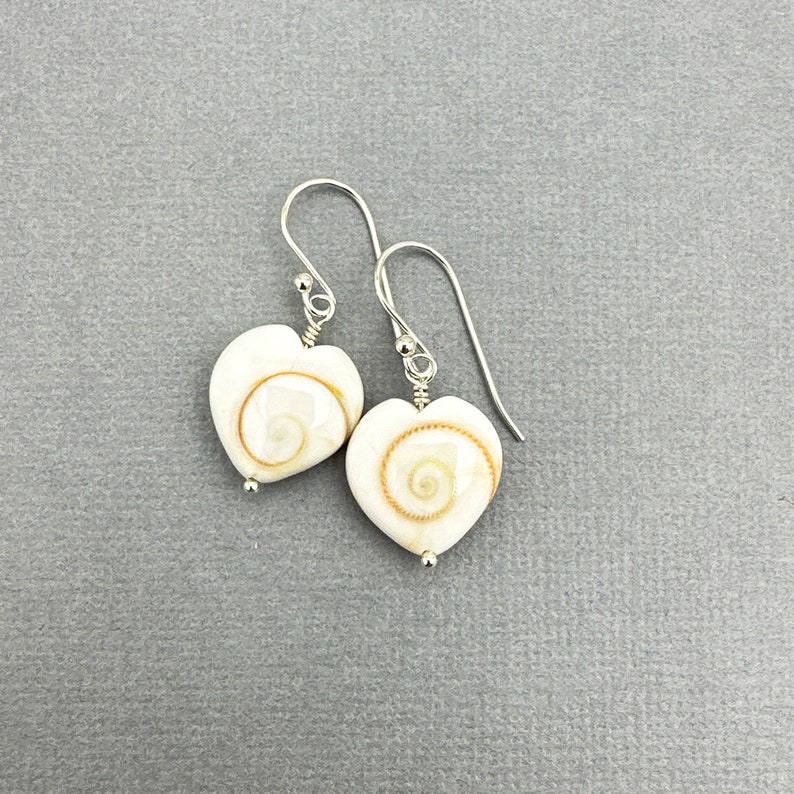 May include: A pair of heart-shaped earrings. Each earring features a white, heart-shaped shell with a spiral design in shades of tan and brown. The earrings have silver-colored hooks and small silver beads.