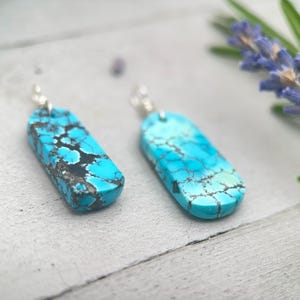 May include: A pair of turquoise earrings with silver hooks. Each earring has a rectangular shape with a polished, crackled surface in shades of blue and black. The earrings are on a light-coloured wooden surface.