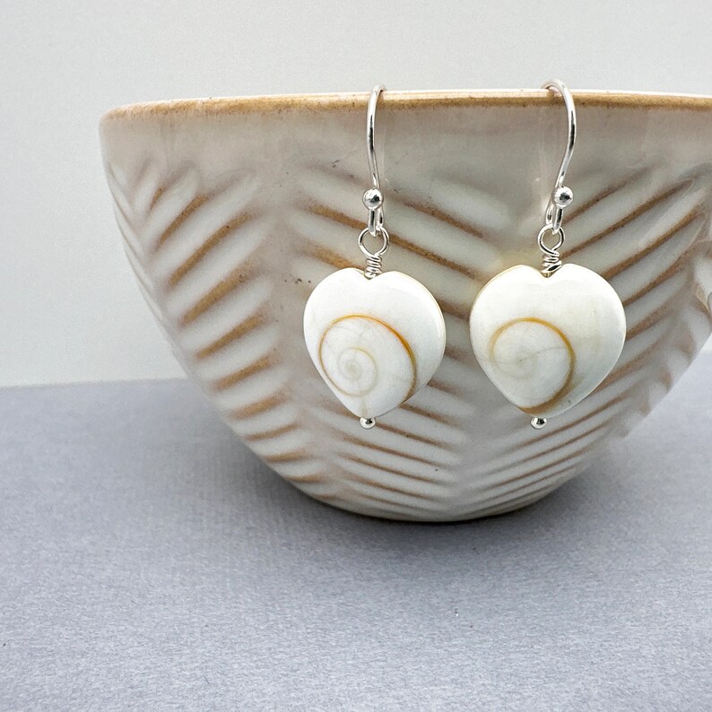 May include: Heart-shaped earrings with a white base and a spiral design in shades of brown and tan. The earrings are suspended from silver-colored hooks. The earrings are displayed in front of a beige bowl.