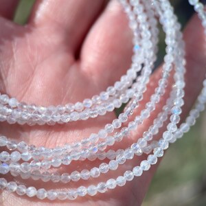 Puede incluir: Una hebra de cuentas de piedra lunar blanca con brillo iridiscente. Las cuentas son peque&ntilde;as y redondas, y la hebra est&aacute; envuelta alrededor de una mano.