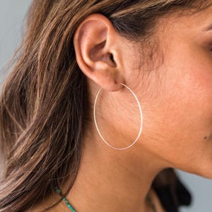 May include: A large, thin, circular hoop earring made of polished gold metal. The earring features a simple, continuous wire design that passes through the earlobe, creating a minimalist and lightweight appearance on the side of the face.