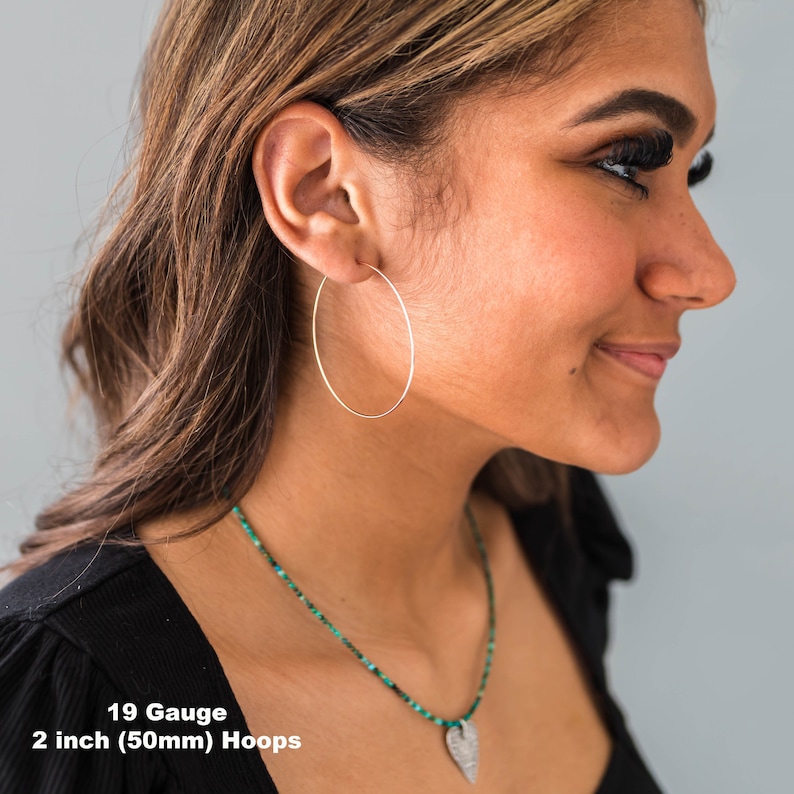 May include: A close-up of a person wearing a pair of large, thin, gold-colored hoop earrings. The earrings are 2 inches (50mm) in diameter and 19 gauge. The text "19 Gauge 2 inch (50mm) Hoops" is visible at the bottom of the image.