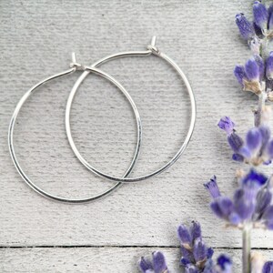 Op de afbeelding: Twee zilveren oorringen met een gehamerde textuur, gepresenteerd op een lichtgrijze houten ondergrond. De oorbellen zijn eenvoudig en elegant. Paarse lavendel takjes zijn op de achtergrond.