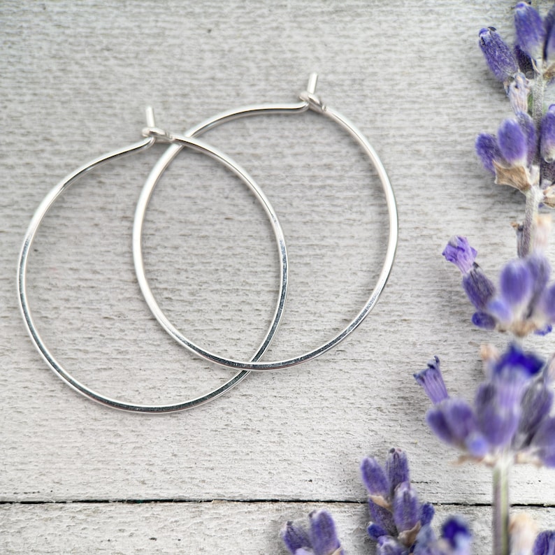 Op de afbeelding: Twee zilveren oorringen met een gehamerde textuur, gepresenteerd op een lichtgrijze houten ondergrond. De oorbellen zijn eenvoudig en elegant. Paarse lavendel takjes zijn op de achtergrond.