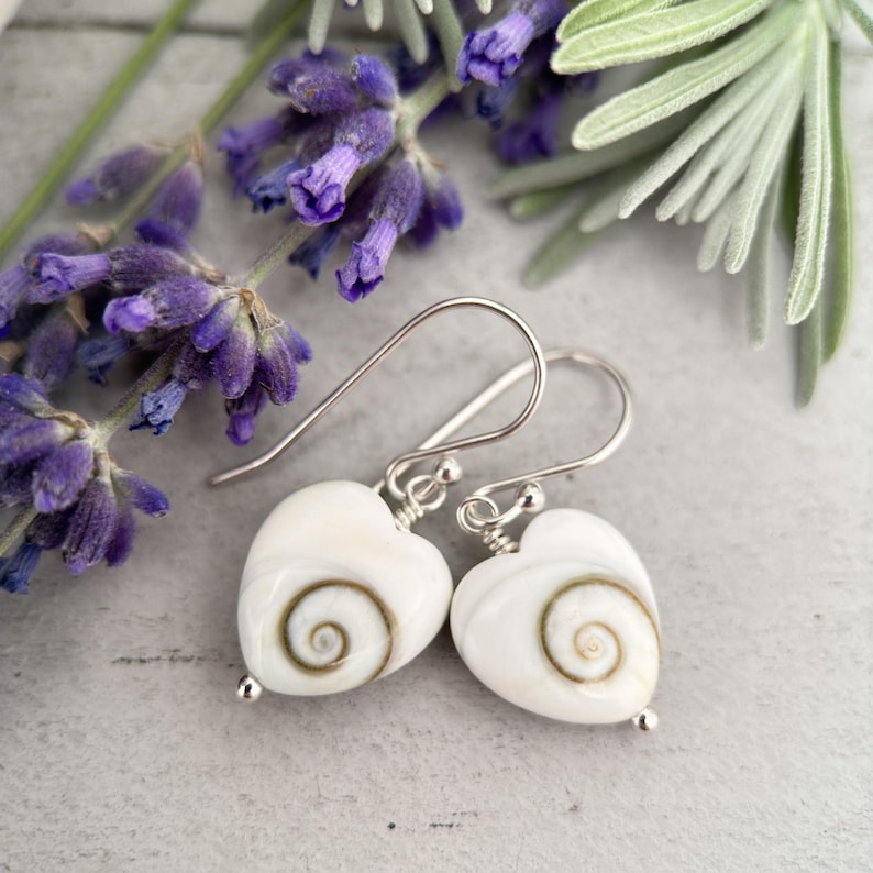 May include: A pair of heart-shaped earrings with a white base and a spiral design in the center. The earrings have silver-colored hooks and small silver beads at the bottom. Lavender flowers and foliage are in the background.