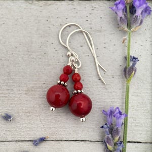 May include: Red beaded earrings with silver hooks. The earrings feature a large, round red bead at the bottom, with smaller red beads and silver accents above. The earrings are displayed on a light gray wooden surface, with sprigs of lavender.