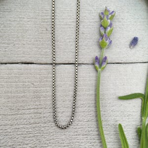 Puede incluir: Un collar de cadena de plata con un diseño delicado de eslabones cuadrados. El collar se muestra sobre una superficie de madera gris claro, junto a una ramita de lavanda con capullos morados y hojas verdes.