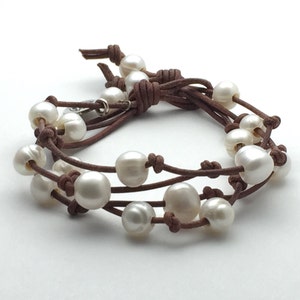 May include: A brown leather bracelet with white pearls. The bracelet is made of multiple strands of leather and has a simple knot closure.