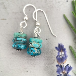 May include: A pair of turquoise and silver earrings. Each earring features three stacked, round turquoise beads with dark veining, suspended from a silver hook. The earrings have a simple, elegant design, perfect for everyday wear. The background is a soft gray.