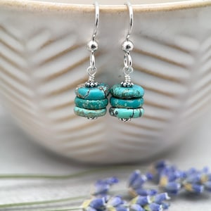 May include: A pair of dangle earrings featuring three stacked turquoise-colored stone discs on each silver-toned hook. The discs display natural veining and are accented with small silver-toned beads at the top and bottom of each stack.