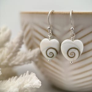 May include: A pair of heart-shaped earrings with a white base and a brown spiral design. The earrings have silver-colored hooks and small silver beads. The earrings are displayed near a white coral and a beige ceramic bowl.