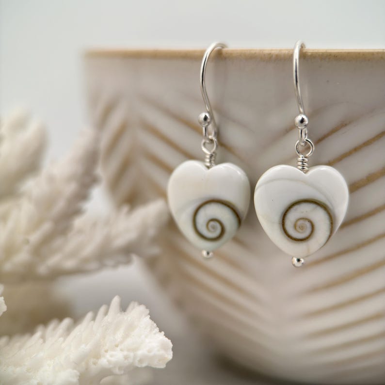May include: A pair of heart-shaped earrings with a white base and a brown spiral design. The earrings have silver-colored hooks and small silver beads. The earrings are displayed near a white coral and a beige ceramic bowl.