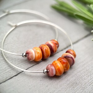 May include: A pair of silver hoop earrings with orange and pink spiny oyster shell beads. The beads are strung along the bottom of the hoops, with a small silver bead separating the shell beads. The earrings are on a light gray wooden surface.