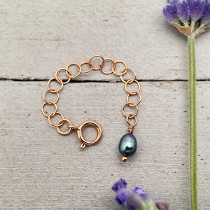 Può includere: Un delicato braccialetto a catena dorata con chiusura, con un singolo pendente di perla blu-verde scuro. Il braccialetto è esposto su una superficie di legno grigio chiaro, con un rametto di lavanda sullo sfondo.