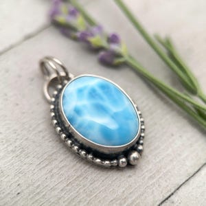 Puede incluir: Un colgante de forma ovalada con una piedra azul claro lisa y patrones de veteado blanco. La piedra est&aacute; engastada en un bisel de plata oxidada oscura, decorado con una textura granulada de cuentas en el borde y una peque&ntilde;a anilla de plata.