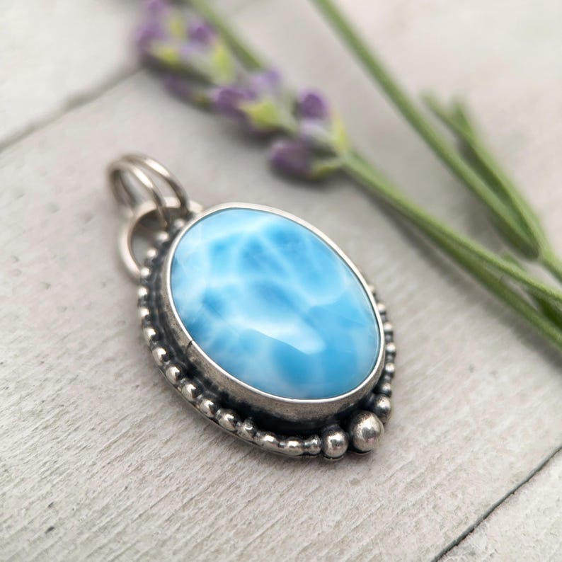 Puede incluir: Un colgante de forma ovalada con una piedra azul claro lisa y patrones de veteado blanco. La piedra est&aacute; engastada en un bisel de plata oxidada oscura, decorado con una textura granulada de cuentas en el borde y una peque&ntilde;a anilla de plata.