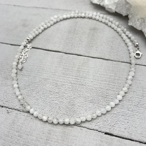 Puede incluir: Un collar de cuentas de piedra lunar blanca con un cierre plateado. El collar est&aacute; sobre una superficie de madera blanca.