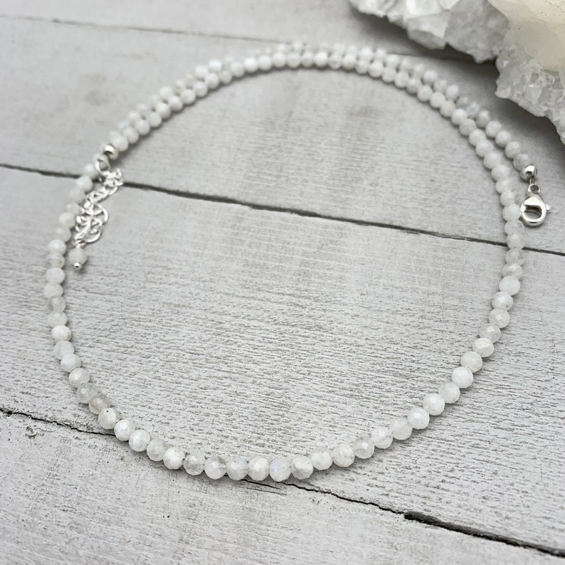 Puede incluir: Un collar de cuentas de piedra lunar blanca con un cierre plateado. El collar est&aacute; sobre una superficie de madera blanca.