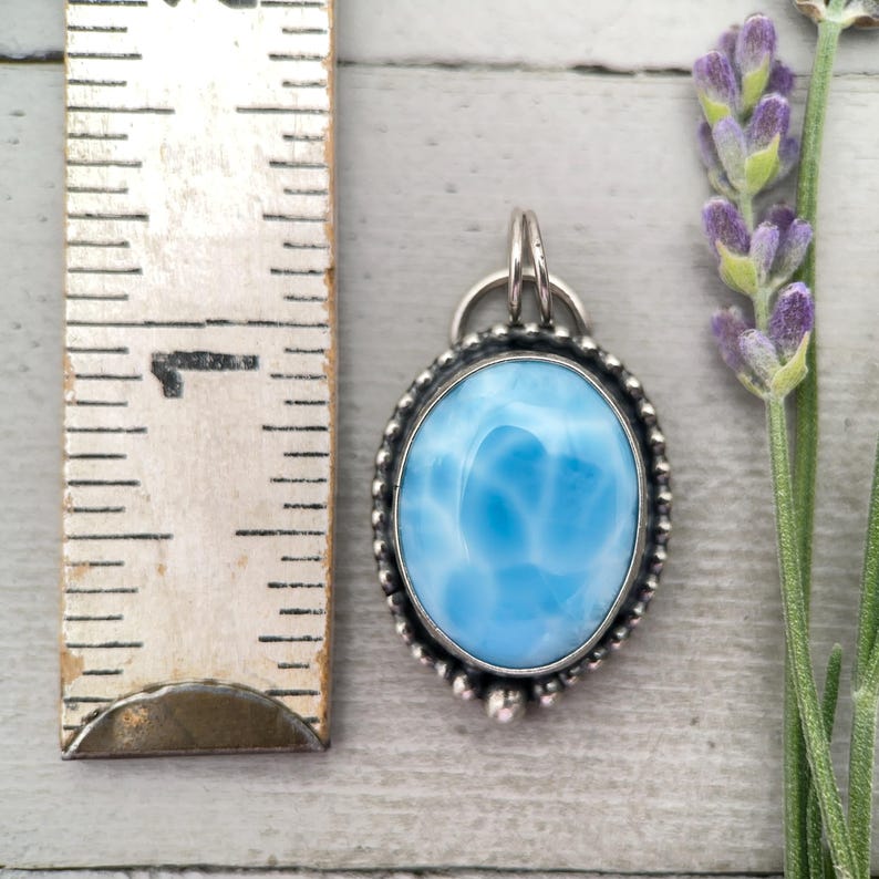 Puede incluir: Un colgante ovalado con una piedra azul claro veteada engastada en un marco de plata con borde de cuentas y una reasa. Una regla colocada junto al colgante muestra que mide aproximadamente 2,5 cm de altura desde la reasa hasta el borde inferior.