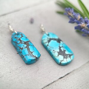 May include: Two turquoise earrings with a rectangular shape and silver hooks. The earrings feature a mottled pattern of black and blue. They are displayed on a light grey surface.