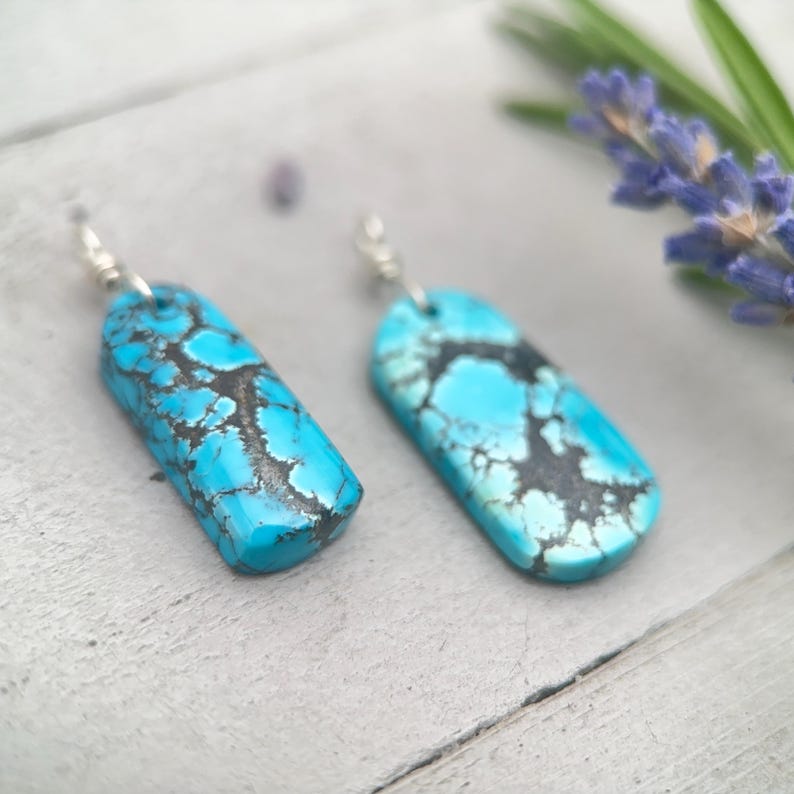 May include: Two turquoise earrings with a rectangular shape and silver hooks. The earrings feature a mottled pattern of black and blue. They are displayed on a light grey surface.