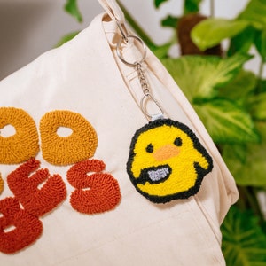 May include: A white canvas tote bag with the words "Good Vibes" embroidered in orange and red yarn. A yellow felt keychain with a black outline and a gray beak is attached to the bag.