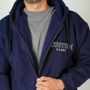May include: A navy blue zip-up hoodie with the word "CUSTOM" and the date "12.6.2023" embroidered in white. The hoodie has a drawstring hood and a full-length zipper. The person is wearing a grey t-shirt underneath.