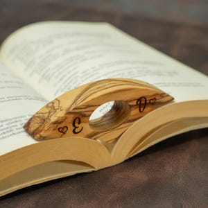 Könnte beinhalten: Ein hölzerner Buchseitenhalter mit natürlichem Holzmaserungsmuster, der auf den Seiten eines offenen Buches liegt. Der Halter hat eine kreisförmige Öffnung und die Buchstaben "E" und "D" mit Herzsymbolen. Ein einzigartiges Accessoire für Bücherliebhaber.