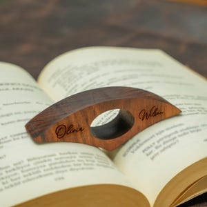 May include: A wooden book page holder with a dark brown finish, featuring an oval opening and the names "Olivia" and "Wilson" engraved on the surface. The holder is resting on the open pages of a book.