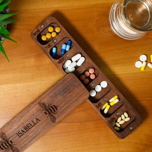 May include: A wooden pill organiser with compartments labelled for each day of the week, filled with various pills. The lid is engraved with a bee and the name "ISABELLA".