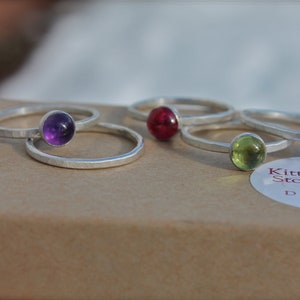 May include: Five silver rings with different colored gemstones. The rings are stacked on top of each other. The gemstones are amethyst, ruby, peridot, and garnet. The rings are on a brown surface with a white label that says "Kitty Stoykovich Designs".